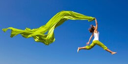 A woman jumps joyfully in the air, holding flowing green fabric against a clear blue sky.