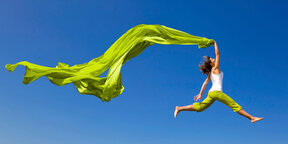 A woman jumps joyfully in the air, holding flowing green fabric against a clear blue sky.