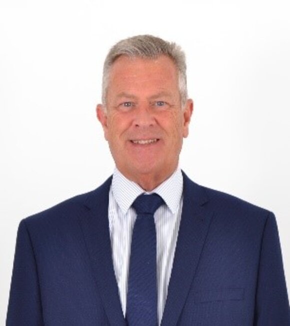A smiling man in a dark suit and tie, with short grey hair, posed against a plain white background.