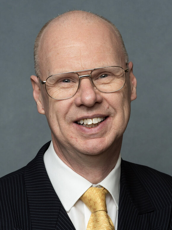 A smiling man in a suit and tie, wearing glasses, stands against a neutral background.