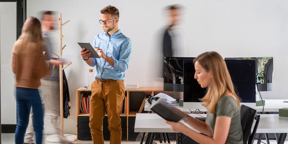 A man using a tablet stands in an office while a woman reads a book nearby, with blurred figures moving in the background.