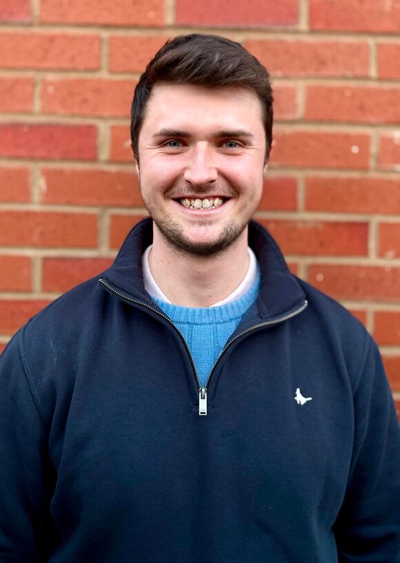 A young man smiling in a dark sweater against a red brick wall.