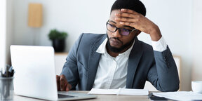 A man in a suit appears stressed while working at a laptop, with a notebook and pen on his desk.